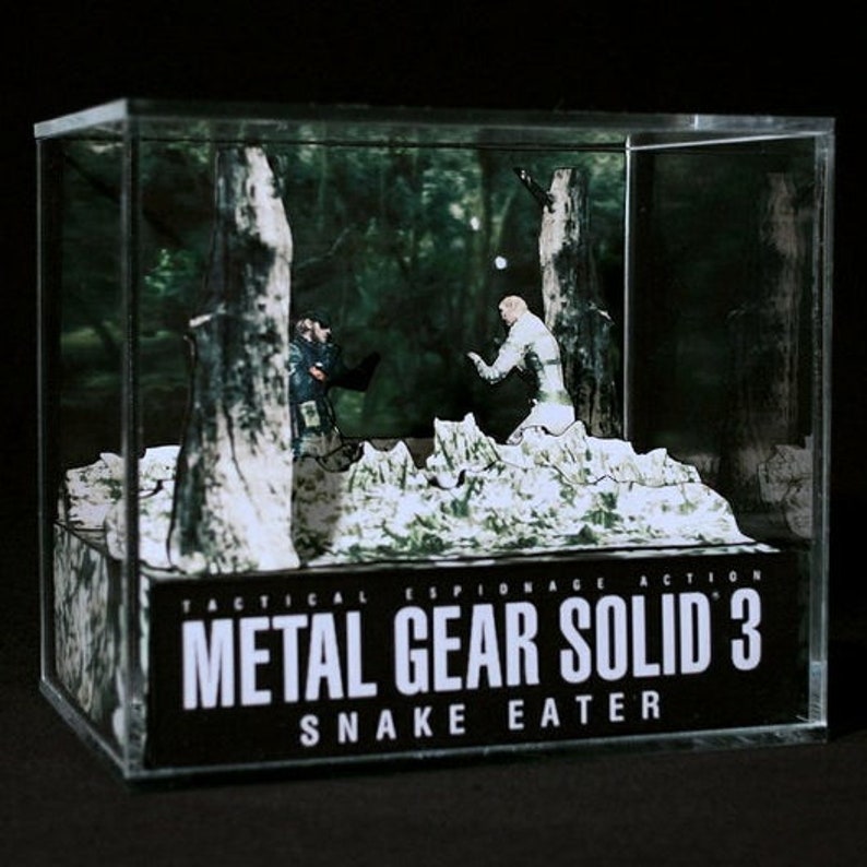 Puede incluir: Una vitrina de pl&aacute;stico transparente con un diorama en su interior que representa una escena del videojuego Metal Gear Solid 3: Snake Eater. El diorama presenta dos figuras, una con un traje de camuflaje y la otra con un uniforme militar, de pie en un entorno de bosque. El texto "Tactical Espionage Action Metal Gear Solid 3 Snake Eater" est&aacute; impreso en la parte frontal de la vitrina.