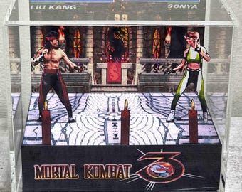 Op de afbeelding: Een doorzichtige acryl displaybox met een 3D-diorama van een Mortal Kombat 3-videogamescène. De scène toont Liu Kang en Sonya Blade tegenover elkaar op een stenen vloer met kaarsen, met de tekst Mortal Kombat 3 op de voorzijde.