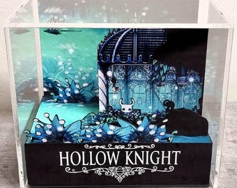 Könnte beinhalten: Eine durchsichtige Acryl-Displaybox mit einem mehrschichtigen Papier-Diorama des Videospiels Hollow Knight. Die Szene zeigt einen blau getönten Wald mit leuchtender Flora und einer zentralen Spielfigur. Der schwarze Sockel trägt den Text HOLLOW KNIGHT.