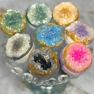 Könnte beinhalten: Eine Auswahl an bunten, geodenförmigen Badekugeln. Jede Badekugel hat eine einzigartige Farbe, darunter Grün, Blau, Gold, Lila und Pink, mit einem goldfarbenen Rand. Sie sind auf einer dekorativen Glas-Kuchenplatte angeordnet.