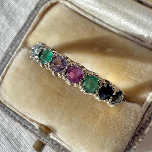 May include: A silver ring set with a line of vibrant gemstones, including green, purple, and pink. The ring is presented in a vintage jewellery box with a cream velvet lining. The ring's design is delicate and elegant.