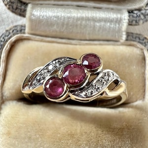 May include: A vintage gold ring with three oval-cut ruby gemstones and small diamond accents. The ring has a swirling design, with the rubies in the center and diamonds along the sides. It's displayed in a velvet-lined jewellery box.