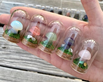 May include: Six small glass jars with cork tops, each containing a miniature mushroom made of different colored gemstones. The mushrooms are displayed on a hand.