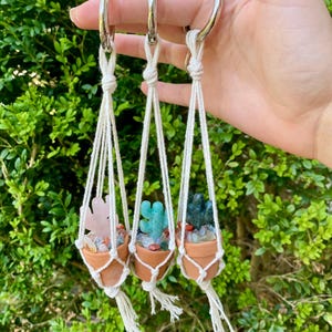 May include: Three small terracotta pots with macrame hangers, each containing a different crystal cactus. The pots are filled with colorful crystals and the cactuses are made of green and pink stones.