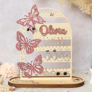 以下が含まれることがあります： ピンクの蝶のデザインと「Olivia」という名前が書かれた木製イヤリングホルダー。ホルダーにはイヤリング用のピンが5列と、ネックレスやブレスレット用の下段の棚があります。
