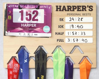May include: A wooden display board with the name "HARPER'S" and personal best times for running events. It features a race bib, medal ribbons, and slots for hanging medals. The board includes times for 5K, 10K, Half, and Full marathons.