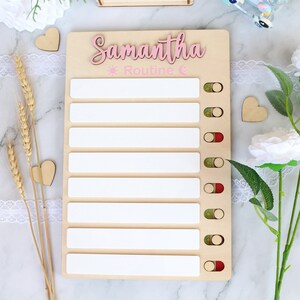 May include: A wooden board with a pink inscription that says "Samantha Routine" with a sun and moon icon. The board has eight white spaces for writing and eight red and green toggle buttons.