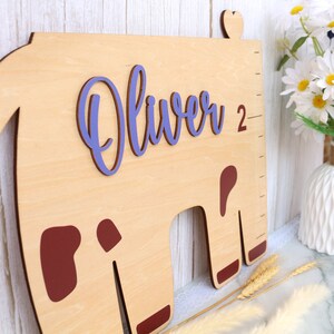 May include: A wooden growth chart shaped like a cow with brown spots. The chart is painted light brown and has a purple name "Oliver" and the number "2" on it.