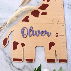 May include: A wooden giraffe growth chart with a purple name cutout that says "Oliver" and a red heart cutout. The giraffe is brown with red spots and has a ruler on the side.