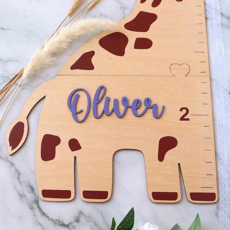 May include: A wooden giraffe growth chart with a purple name cutout that says "Oliver" and a red heart cutout. The giraffe is brown with red spots and has a ruler on the side.