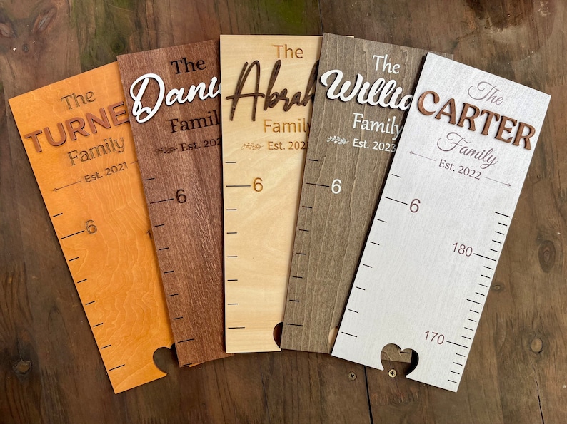May include: Five wooden growth charts with different wood stains and personalised family names. The charts feature a heart cutout at the bottom and a ruler with markings up to 1.8 metres. The names on the charts are "The Turne Family Est. 2021", "The Dani Family Est. 202", "The Abrah Family Est. 202", "The Willia Family Est. 2023", and "The Carter Family Est. 2022".