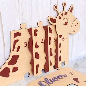 May include: Wooden giraffe growth chart with brown giraffe spots and numbers 2 through 6. The chart is made of light wood and has a purple nameplate that says "Oliver" at the bottom.