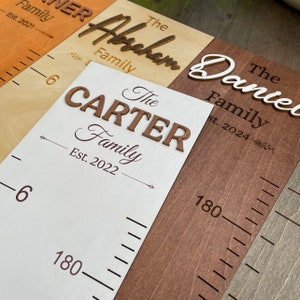 May include: Three wooden growth charts with different family names and establishment dates. The charts are made of different wood tones, including light brown, dark brown, and white. The names are "The Carter Family Est. 2022", "The Abraham Family", and "The Daniel Family Est. 2024".