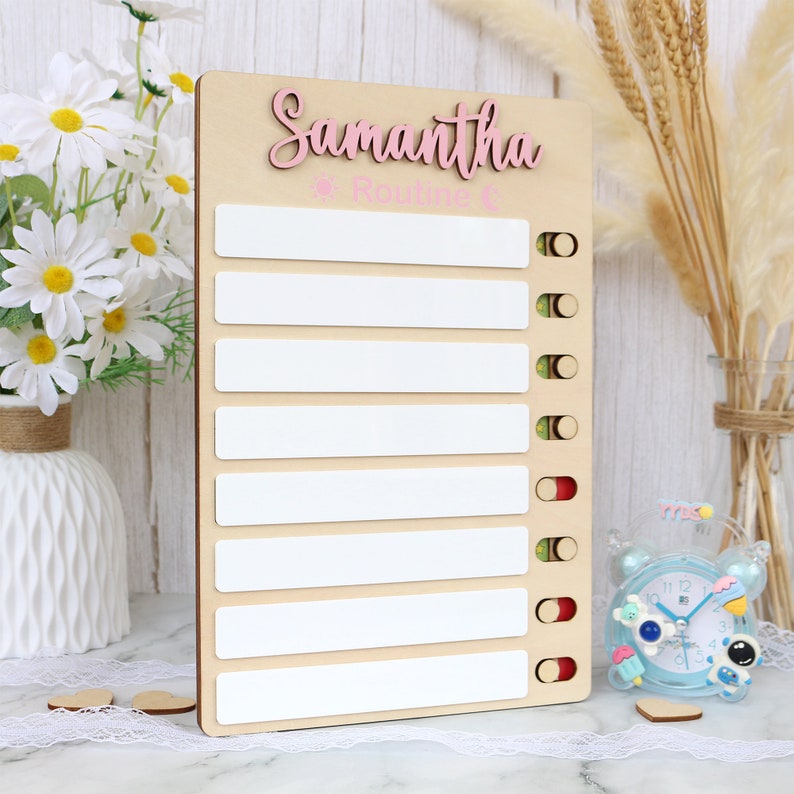 May include: A wooden board with a white background and a pink inscription that reads "Samantha's Routine". The board has eight white spaces for writing with red, green, and yellow circles next to each space.