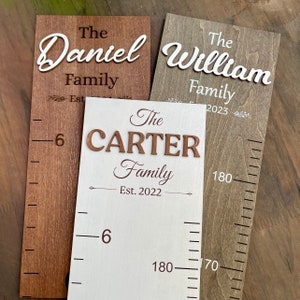 May include: Three wooden growth charts with white and brown paint. The charts feature the names "The Daniel Family", "The William Family", and "The Carter Family" with the year established. The charts have a ruler on the side with measurements in centimetres.
