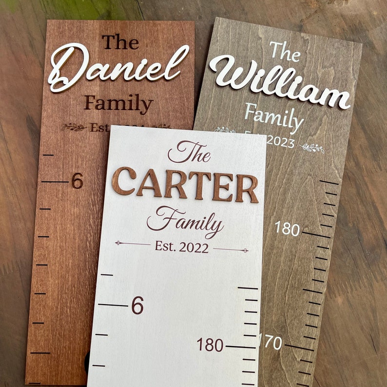 May include: Three wooden growth charts with white and brown paint. The charts feature the names "The Daniel Family", "The William Family", and "The Carter Family" with the year established. The charts have a ruler on the side with measurements in centimetres.
