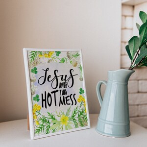 Puede incluir: Impresi&oacute;n enmarcada en blanco con un fondo de acuarela de hojas verdes y flores amarillas. La impresi&oacute;n dice "Jesus loves this hot mess" en letras cursivas negras.