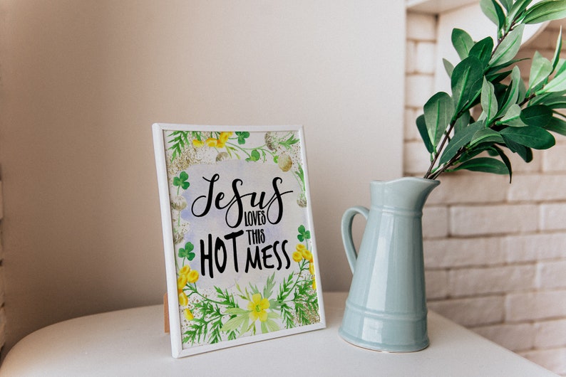 Puede incluir: Impresi&oacute;n enmarcada en blanco con un fondo de acuarela de hojas verdes y flores amarillas. La impresi&oacute;n dice "Jesus loves this hot mess" en letras cursivas negras.