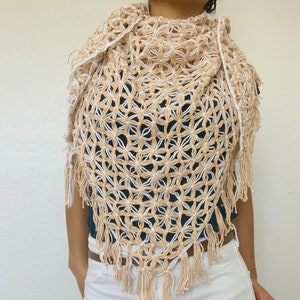 May include: A crocheted shawl with a floral pattern in beige and white. The shawl has fringe along the bottom edge.