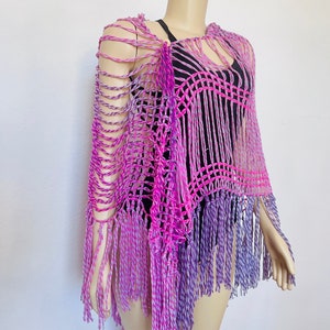 May include: A crocheted poncho with a pink and purple ombre design and fringe. The poncho is made with a loose, open stitch and has a relaxed fit.
