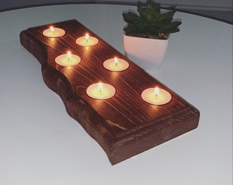 Pode incluir: Um porta-velas de madeira rústico com seis porta-velas para velas votivas. O porta-velas é feito de madeira marrom escura com um acabamento natural e toscamente talhado. As velas estão acesas e brilhando.