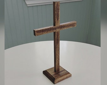 May include: A wooden cross with a dark brown finish. The cross is standing upright on a square base. The horizontal beam of the cross is centered. The cross is displayed on a white table.
