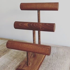 May include: A wooden jewelry stand with three tiers for displaying bracelets. The stand is made of dark brown wood and has a rectangular base.
