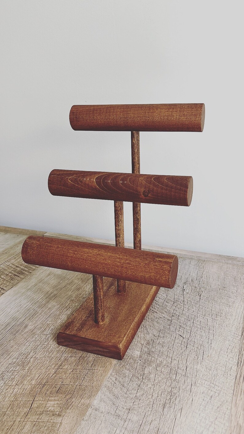 May include: A wooden jewelry stand with three tiers for displaying bracelets. The stand is made of dark brown wood and has a rectangular base.