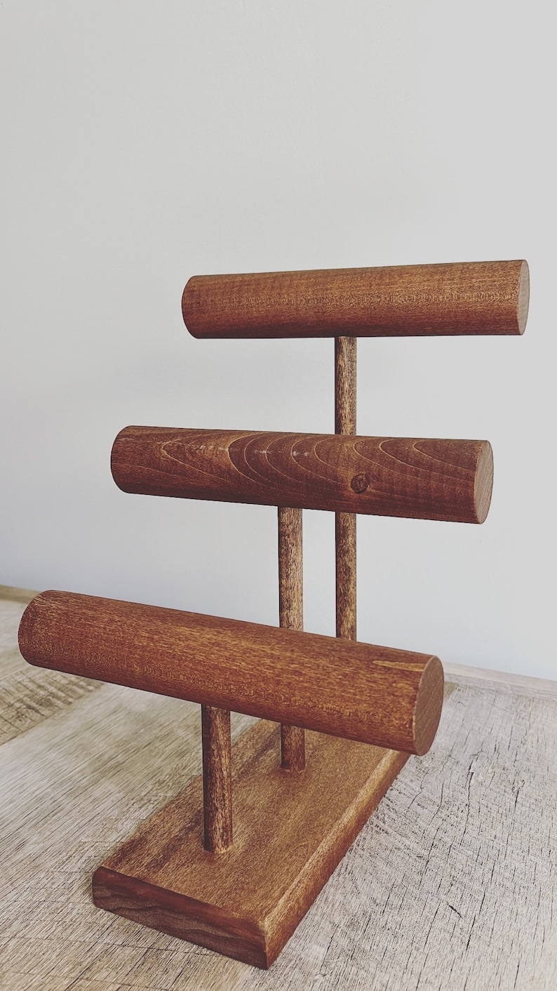 May include: A wooden jewelry stand with three tiers for displaying bracelets. The stand is made of dark brown wood and has a rectangular base.