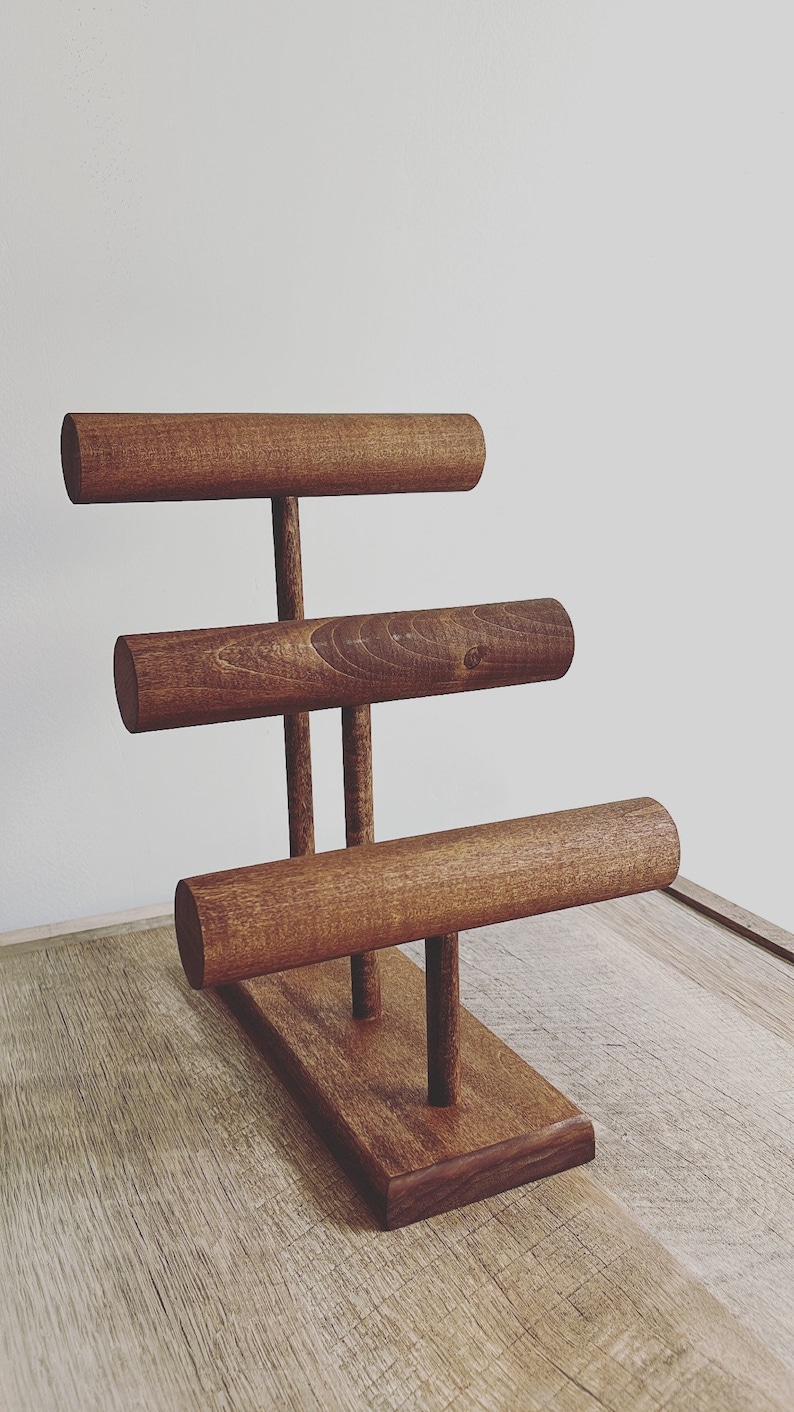 May include: A wooden jewelry stand with three tiers for displaying bracelets. The stand is made of dark brown wood and has a rectangular base.