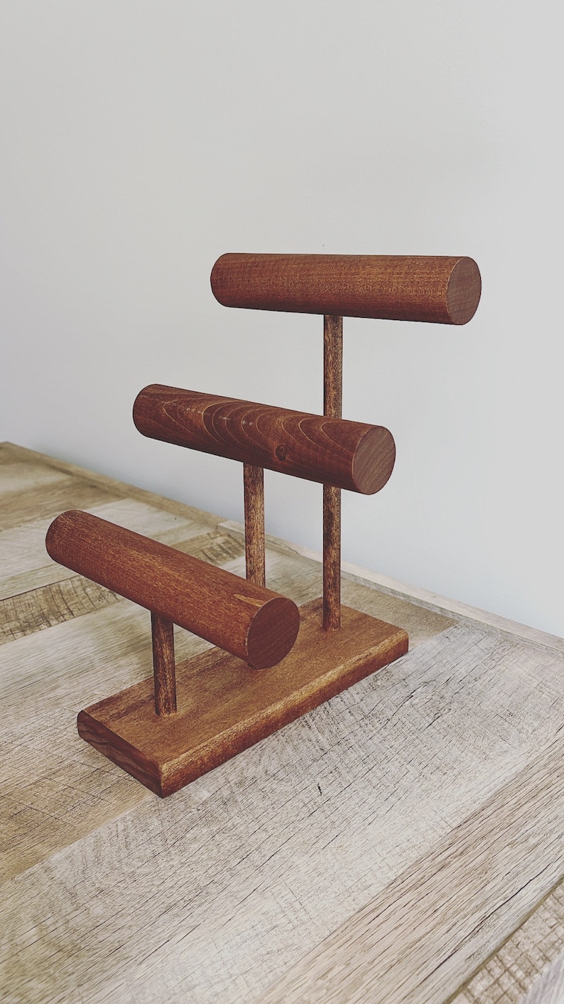 May include: A wooden jewelry stand with three tiers for displaying bracelets. The stand is made of dark brown wood and has a simple, modern design.