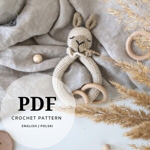 May include: Crochet pattern PDF for a beige bunny rattle toy with wooden rings. The pattern is available in English and Polish.