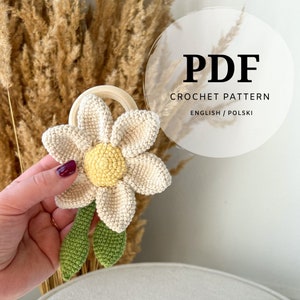 Op de afbeelding: Haakpatroon PDF voor een witte madeliefjesbloem met een geel hart en groene bladeren bevestigd aan een houten ring. Het patroon is beschikbaar in het Engels en Pools.