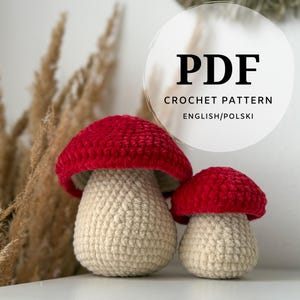 May include: Two crocheted mushroom figures. The mushroom caps are a vibrant red, and the stems are a creamy beige. The text "PDF CROCHET PATTERN ENGLISH/POLSKI" is displayed in a white circle.