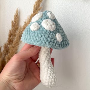 May include: A plush crochet mushroom featuring a soft, textured light blue cap adorned with several rounded white spots. The stem of the mushroom is crafted from a solid white yarn, providing a contrasting base to the colorful, patterned cap.