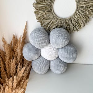 May include: A fabric flower decoration with a light grey and white colour scheme. The flower is made up of six light grey spheres around a white central sphere. A circular wall hanging and dried plants are visible in the background.