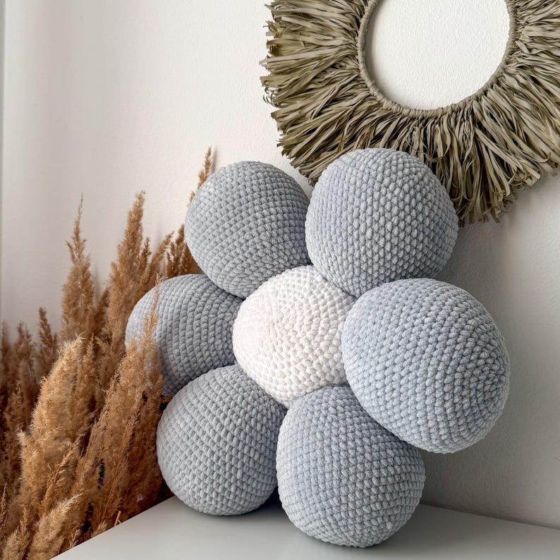 May include: A collection of handmade, round, plush cushions in shades of grey and white. The cushions are arranged in a flower-like pattern. A decorative wall hanging and dried plants are in the background.