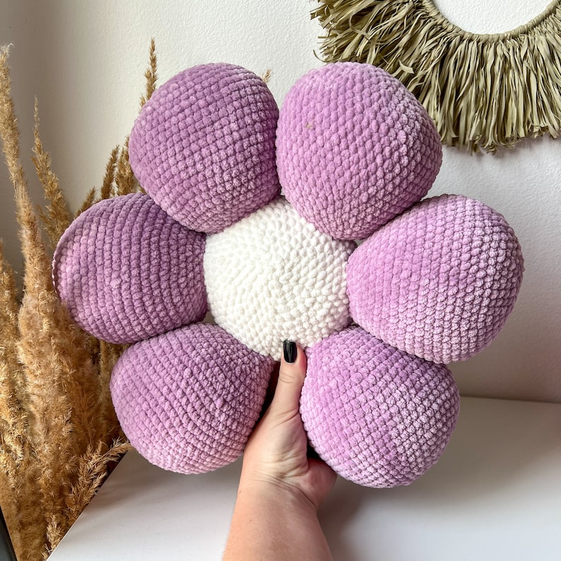 May include: A flower-shaped cushion with a white central section and lavender petals. The cushion is made from a soft, textured fabric. The item is held by a hand. This is a decorative item.