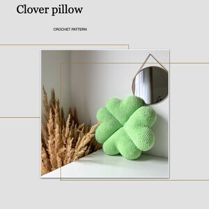 May include: A green crocheted clover-shaped pillow on a white surface. The pillow is in focus, and the background is blurred.