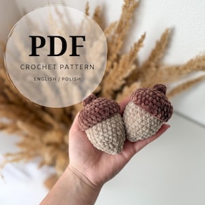 May include: Two handmade crochet acorns in shades of brown and beige are held in a hand. The acorns have a textured, plush appearance. The image includes text that reads "PDF CROCHET PATTERN ENGLISH / POLISH".