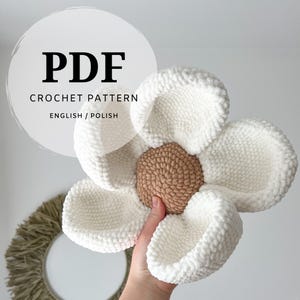 May include: A crochet pattern PDF document featuring text that reads PDF CROCHET PATTERN ENGLISH / POLISH. The image displays a plush, handmade flower with five large, rounded white petals and a textured, circular tan center made of yarn.