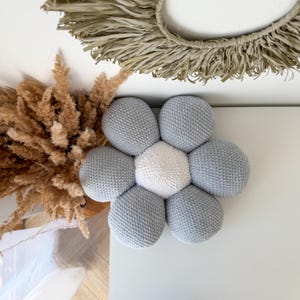 May include: A handmade, grey and white flower-shaped cushion. The cushion has a central white hexagon surrounded by six grey, spherical petals. The cushion is placed on a white surface, with dried plants and a woven wall hanging in the background.