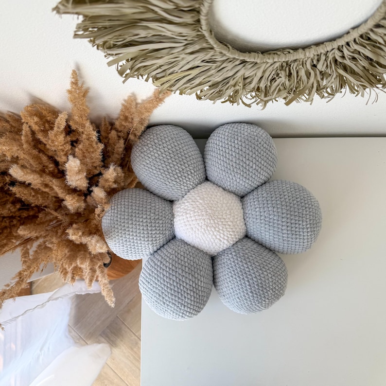 May include: A handmade, grey and white flower-shaped cushion. The cushion has a central white hexagon surrounded by six grey, spherical petals. The cushion is placed on a white surface, with dried plants and a woven wall hanging in the background.