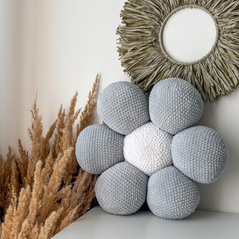 May include: A flower-shaped cushion with a white central section and light grey petals. The cushion is made from a soft, textured material. A decorative wall hanging and dried plants are in the background.