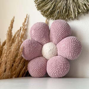 Op de afbeelding: Een groot, bloemvormig kussen in zachtroze en wit. De bloemblaadjes zijn lichtroze en het midden is wit. Het kussen is gemaakt van een getextureerd, gebreid materiaal en wordt waarschijnlijk als decoratief item gebruikt.