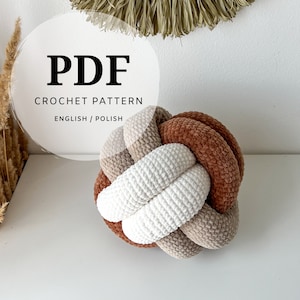 May include: A crocheted knot pillow in shades of white, beige, and brown. The pillow is made of soft, textured yarn and has a unique, knotted design. The image also includes the text "PDF CROCHET PATTERN ENGLISH / POLISH".