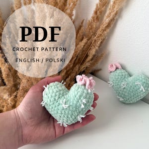Może przedstawiać: Dokument ze wzorem szydełkowym w formacie PDF zatytułowany PDF CROCHET PATTERN ENGLISH / POLSKI przedstawiający dwa pluszowe kaktusy w kształcie serca. Każdy kaktus jest jasny z różowym kwiatkiem i białymi szwami imitującymi kolce.