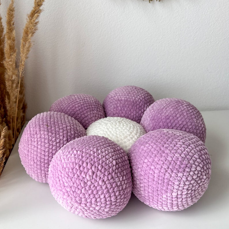 May include: A collection of eight spherical, knitted items. Seven are a pale lilac colour, and one is white. The objects have a textured, plush appearance. The arrangement suggests a decorative or craft item, possibly for home decor or a child's toy.
