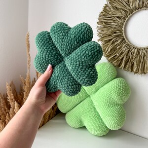 May include: Two green crocheted four-leaf clover pillows. One pillow is held in a hand, the other is on a white surface.