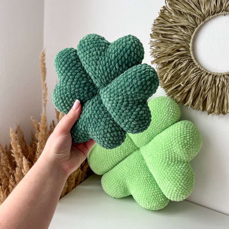 May include: Two green crocheted four-leaf clover pillows. One pillow is held in a hand, the other is on a white surface.